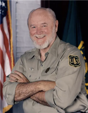 A smiling bearded man with his arms crossed over this Forest Service uniform. 